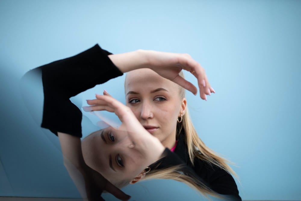 Femeie tânără blondă pe fundal albastru, cu propria reflexie în oglindă dedesubt și brațele ridicate la nivelul feței